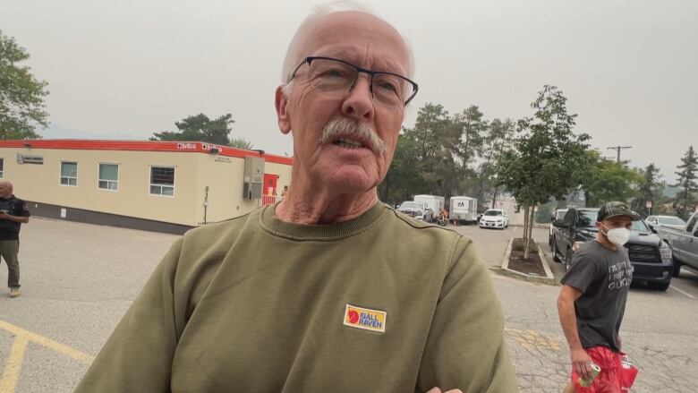 A man with glasses and a grey moustache wears a green T-shirt and speaks.