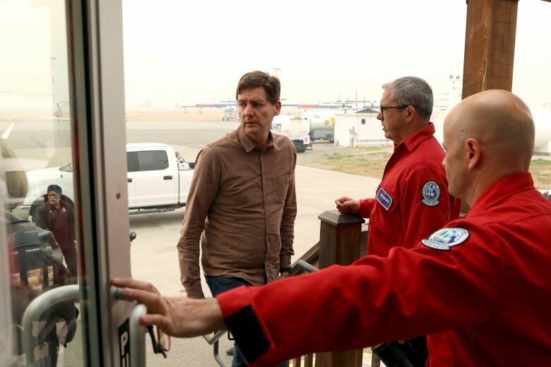 A tall white man wearing a brown shirt is greeted by two men wearing red uniforms.