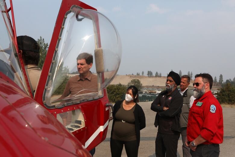 A gaggle of people stare at a red helicopter. One of them, a woman, is wearing a respirator-style mask, while another man is wearing a red uniform.