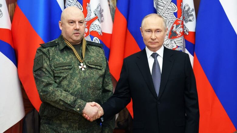 A man in a suit shakes hands with a man in a military uniform who is wearing a medal around his neck.