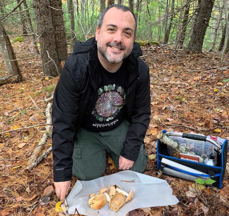 A man with very short dark hair, greying short moustache and goatee, kneels on the forest floor wearing a dark hooded sweatshirt and dark cargo pants. There are several mushrooms on a piece of fabric in front of him, and a toolbox nearby.