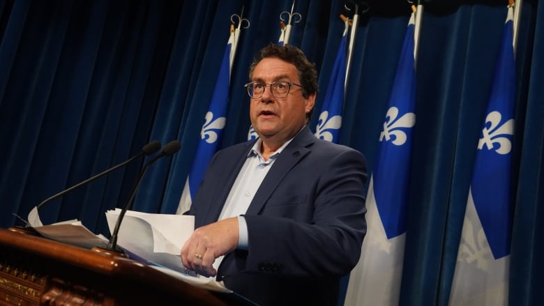 A man speaks at a podium in front of Quebec flags. 