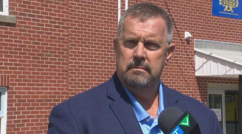 Man with short grey-brown beard and moustache wearing blue suit jacket standing outside brick building.