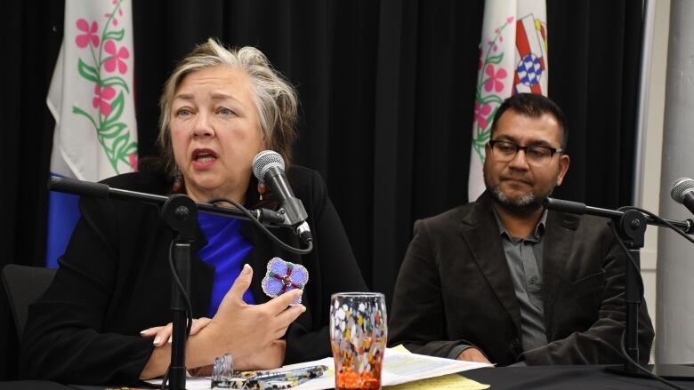 A woman sits at a table speaking into a microphone, while a man sits beside her, looking on.