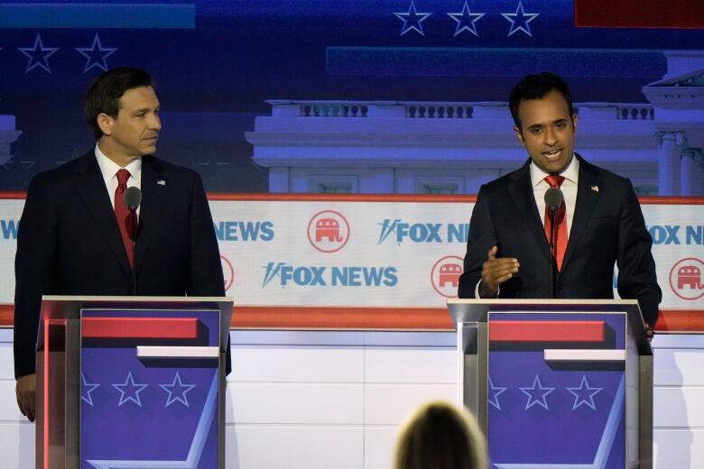 2 men both in dark suits and a red tie stand behind podiums speaking