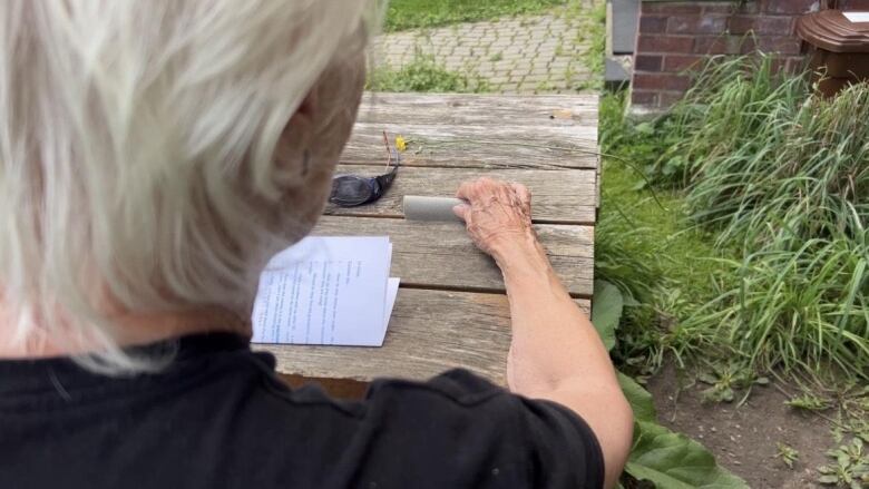 From behind the shoulder, an older woman sits outside at a garden table.