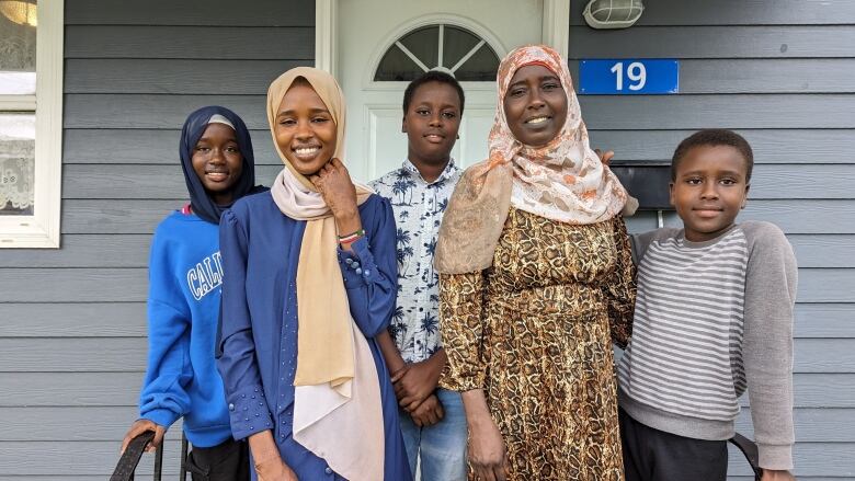 Two young women, two young men and an older woman pose on the stairwell in front of a house. 