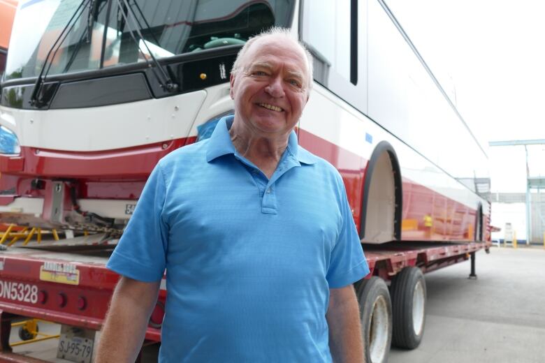 A man with grey hair stands smiling in front of the shell of a red and white transit bus.