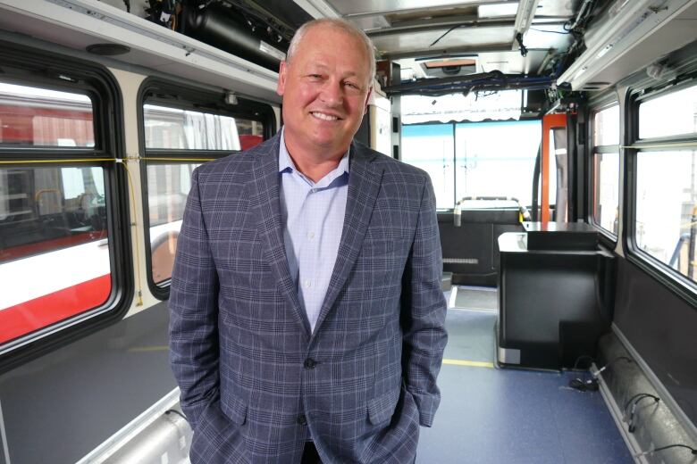 A man in a suit coat stands smiling on board an empty, partially-constructed transit bus 