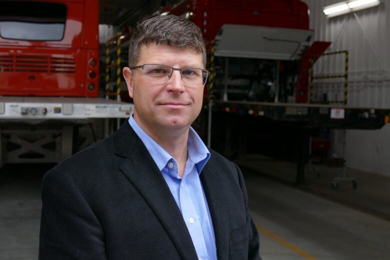 A middle-aged man smiles in front of the bay door to the factory, two partly-built buses behind him.