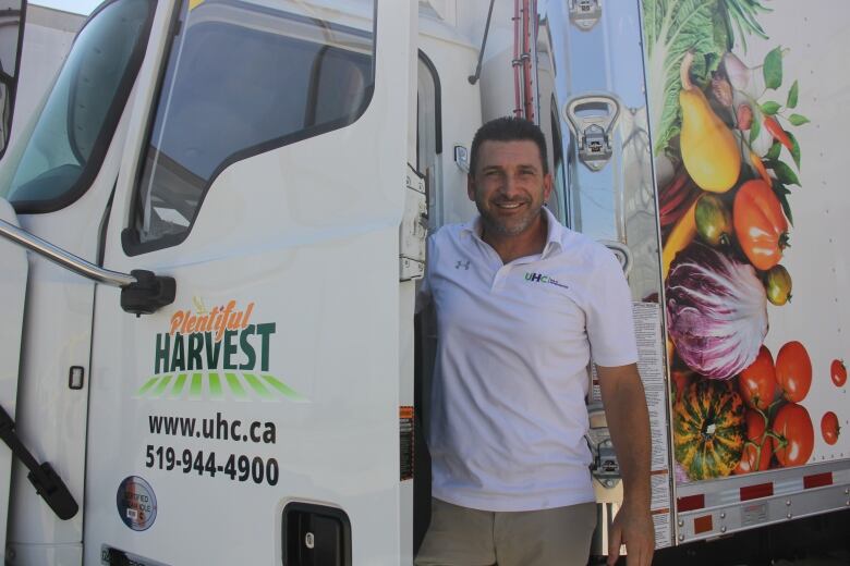 A man in a white polo shirt stands in front of a large truck with vegetables on the side. 