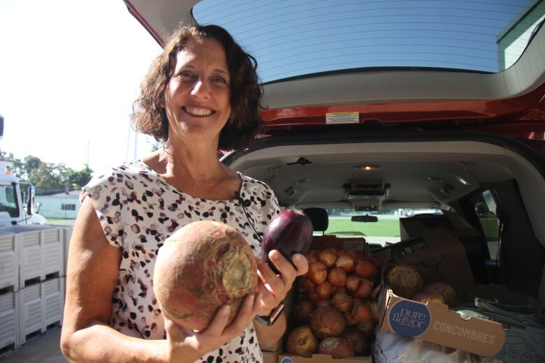 A woman stands in front of an open van holding vegetables. 