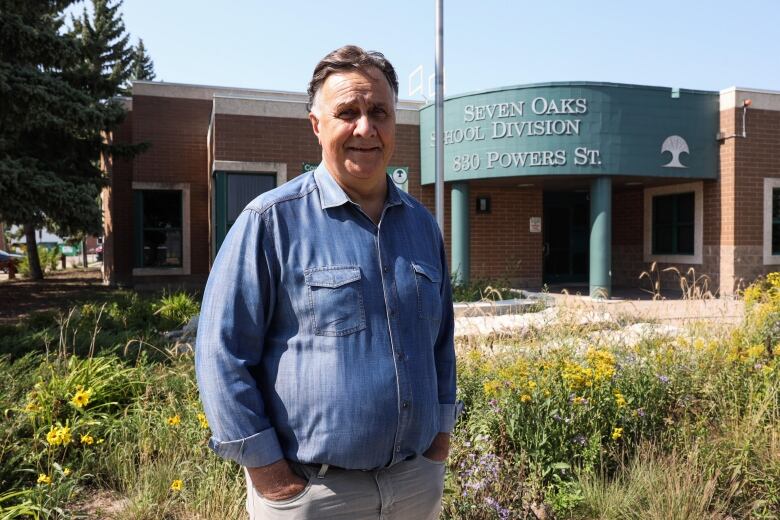 A man stands outside in front of flowers and a building with a green entranceway that called Seven Oaks School Division. 