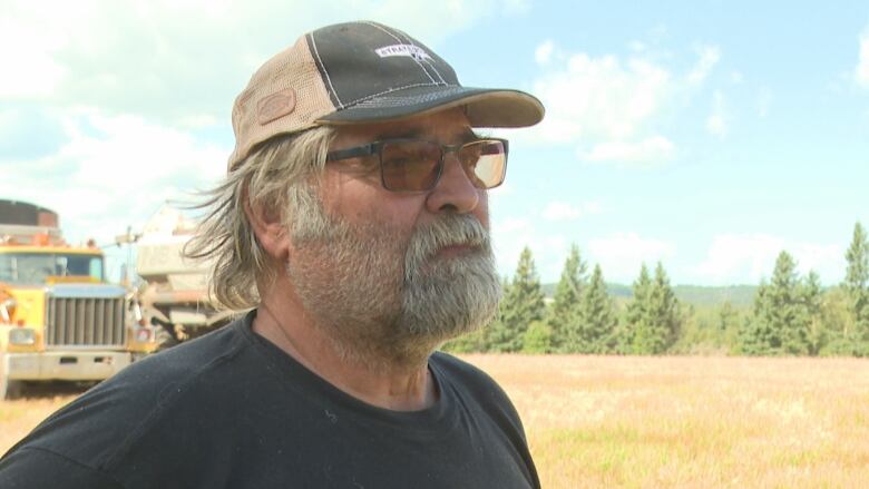 Bearded man in ball cap stands in a field, with a farm truck behind him. 