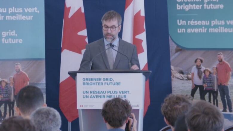 A man with a beard speaks at a podium with flags behind him.