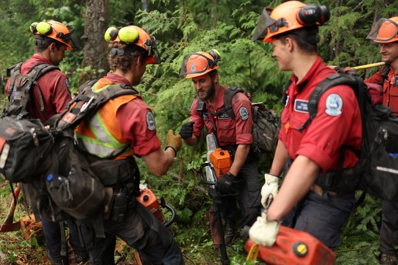 Five young firefighters, dressed in red shirts and orange hardhats with heavy backpacks, stand in a huddle as they play rock-paper-scissors.