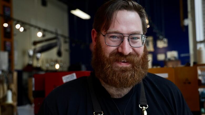 A young man in glasses with a bushy red beard and wearing a dark blue shirt smiles at the camera with his shop blurred out in the background.