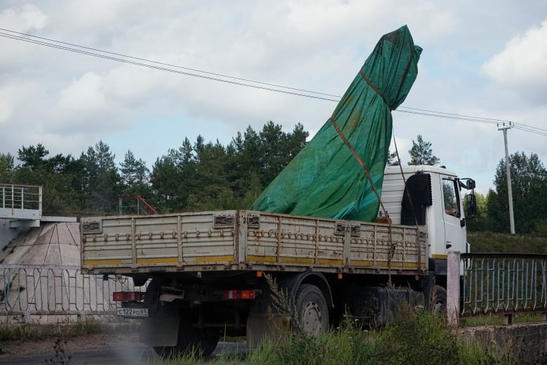 A flatbed truck is shown transporting a very large item, covered and wrapped in a green tarpaulin.