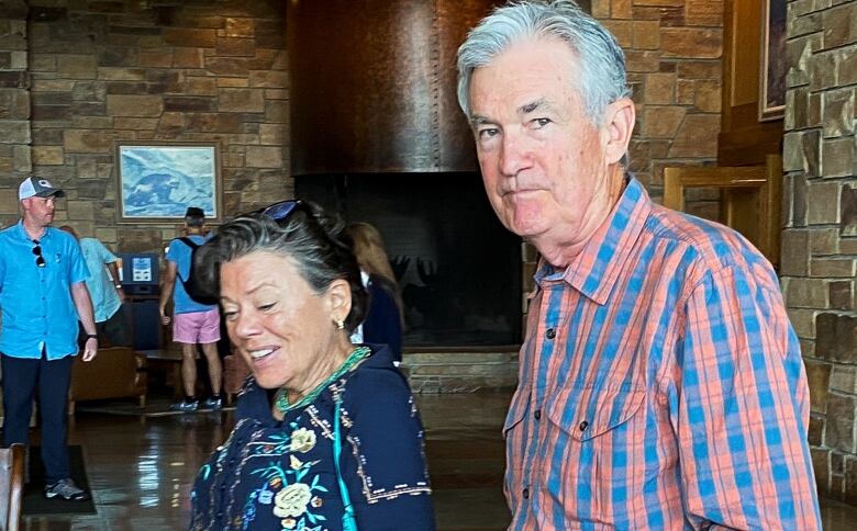 A white-haired man wearing a blue-and-pink checked shirt stands beside a woman with brown-grey hair who's wearing a floral navy top.