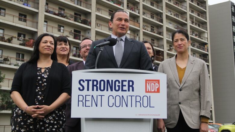 A man stands at a podium, backed by several of his party's candidates. An apartment complex is visible in the background.