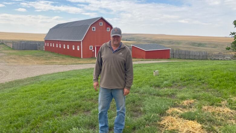 A man in a ballcap and blue jeans stands in a field in front of a red barn.