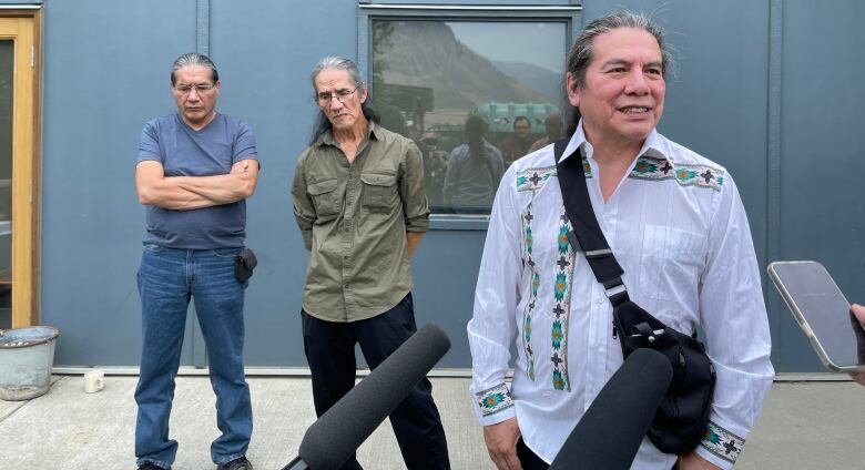 Three Indigenous men stand speaking to microphones.