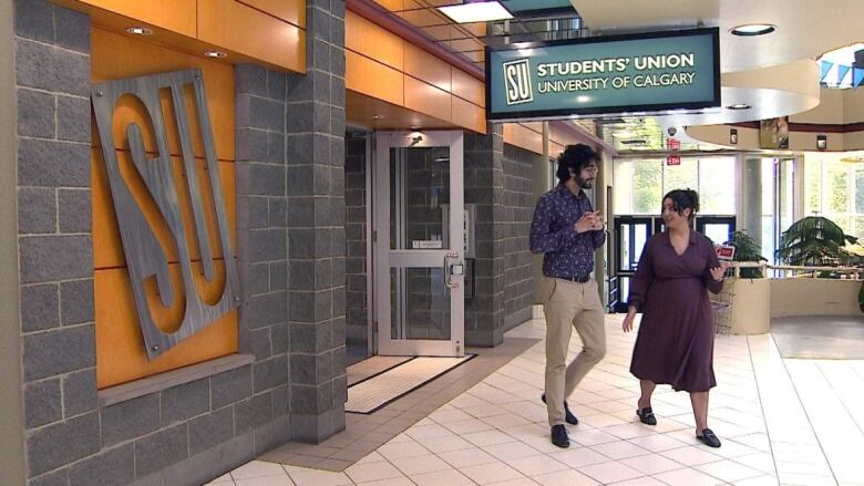 Two people, one woman and one man, walk out of a door with a sign overhead that reads 