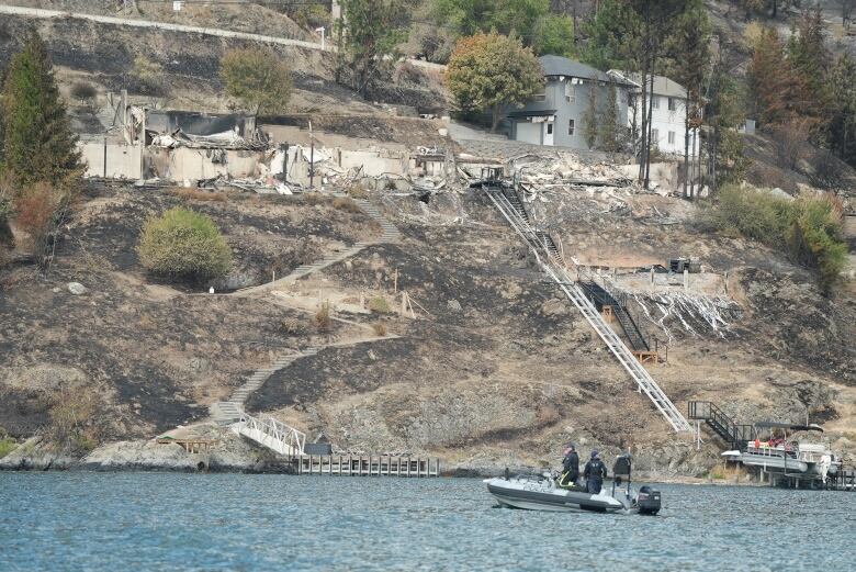 An RCMP boat is seen on a lake. In the foreground is a hilly area with debris and destroyed houses.