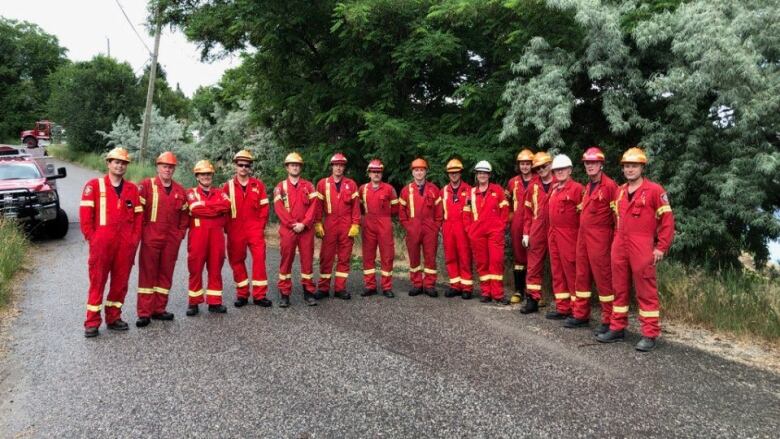 A large group of 15 firefighters stand side by side on the road in red fire fighting outfits. 