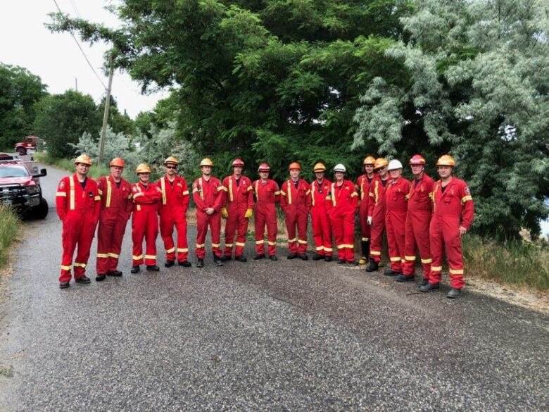 A large group of 15 firefighters stand side by side on the road in red fire fighting outfits. 