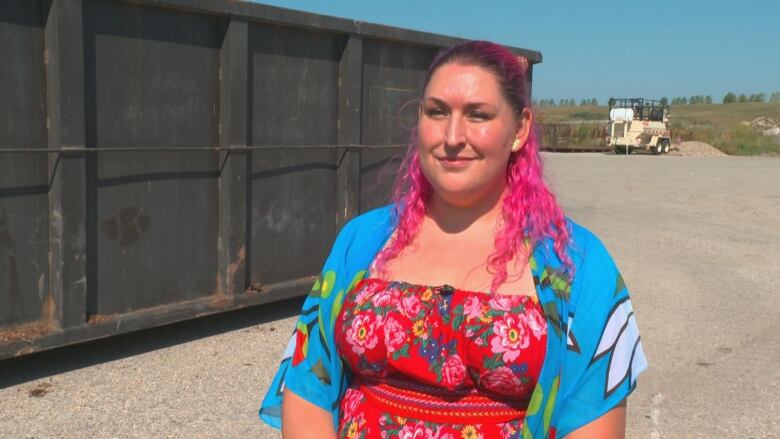 A woman with bright pink hair stands in front of a large dumpster. 