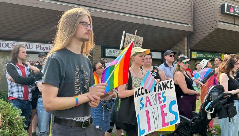 A group of people are standing on a lawn in front of an office building. They are holding pride flags and signs.