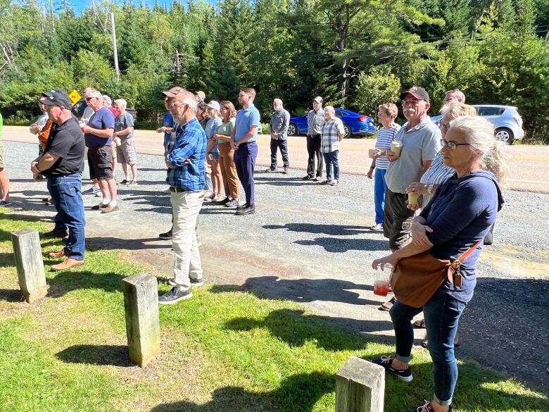 About two dozen people stand on grass looking off camera. They are standing on a roadside and there are trees in the background. 