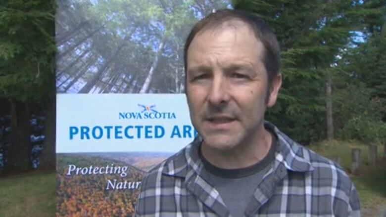 A man in a plaid shirt stands in front of a line of trees and a sign that says Nova Scotia Protected Area, protecting nature.