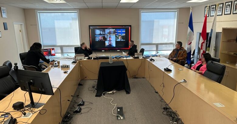 A few people sit in a meeting room, with a video screen at the front of the room.