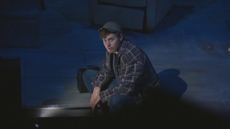 A young man in a plaid coat and old baseball cap looks up wistfully toward the camera while sitting on a darkened stage. 