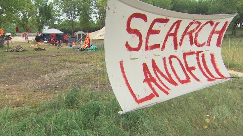 Tents are set up on grass and next to trees. A banner, strung between two trees, says Search Landfills.