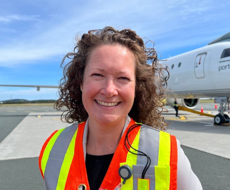 A middle-aged woman smiles. She wears a safety vest and stands in front of an airplane.