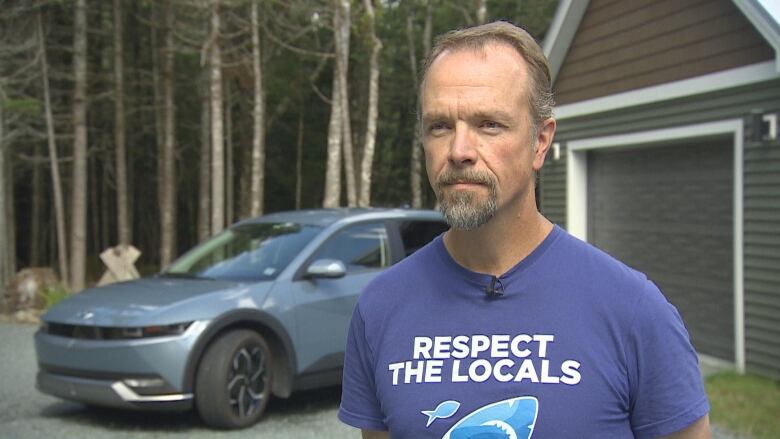 A man with a beard and moustache is show in a blue shirt that reads 'respect the locals'. He is standing against a backdrop of his Hyundai electric vehicle parked near his Nova Scotia home.