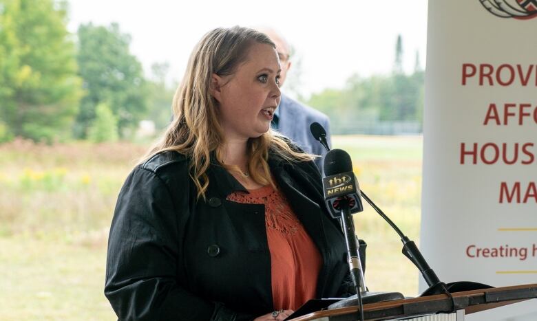 A woman with long blonde hair speaks into a microphone outside.