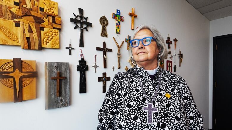 A woman wearing glasses stands in front of a wall of crosses. 