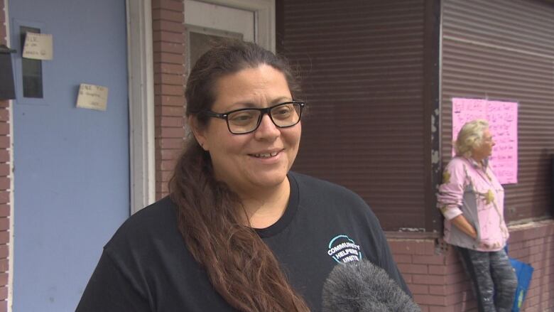 A woman with long dark hair and glass in a black t-shirt speaks with media in front of a brown brick building.