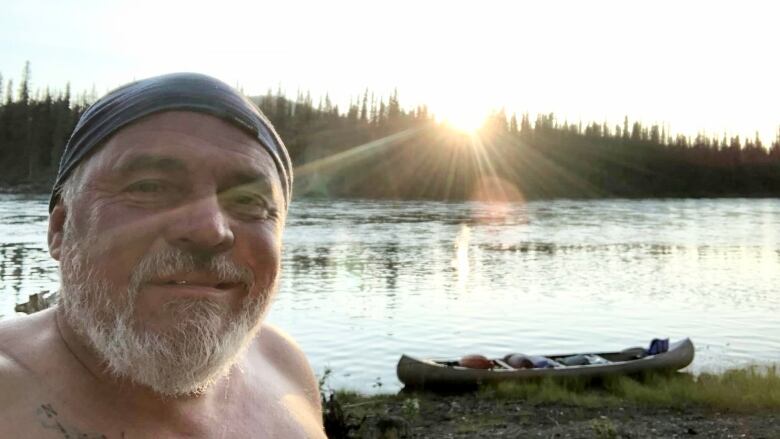 A shirtless man takes a selfie in front of his canoe on the shores of the Yukon River.