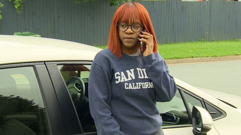 A woman speaks on her phone while standing next to a white car.