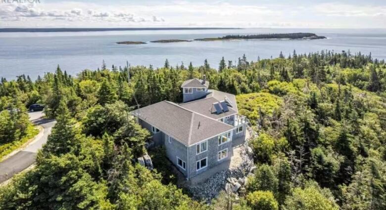 A large grey cedar-shake home is seen perched on a coastal area, surrounded by green trees and a rocky coast with water in the distance.