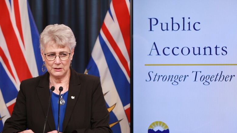 A woman in a black suit speaks on a podium next to a sign that reads 'Public Accounts Stronger Together.'