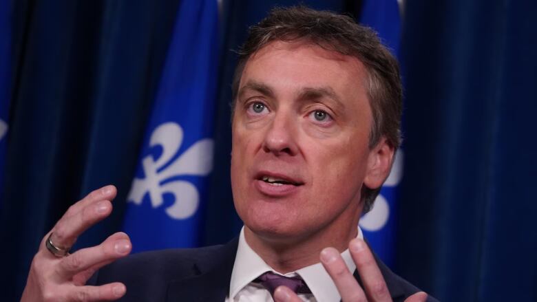 A man in a suit speaks in front of a Quebec flag. 