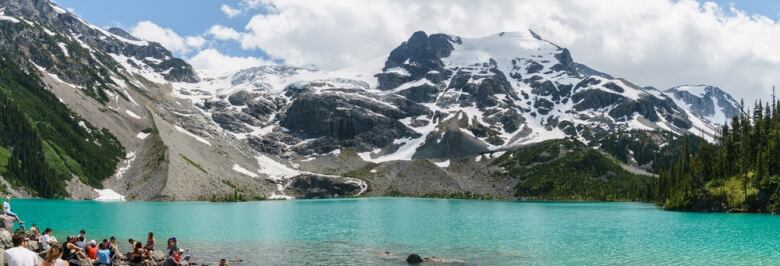 A panorama of a scenic mountain lake with people to the bottom-left.