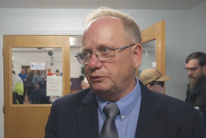 A man with glasses wearing a suit and tie. Two people converse behind him