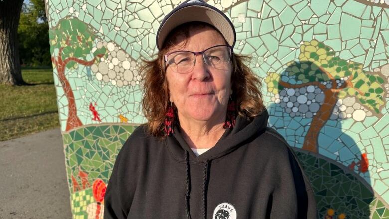 A woman in a black hoodie and ball cap stands in front of a wall.